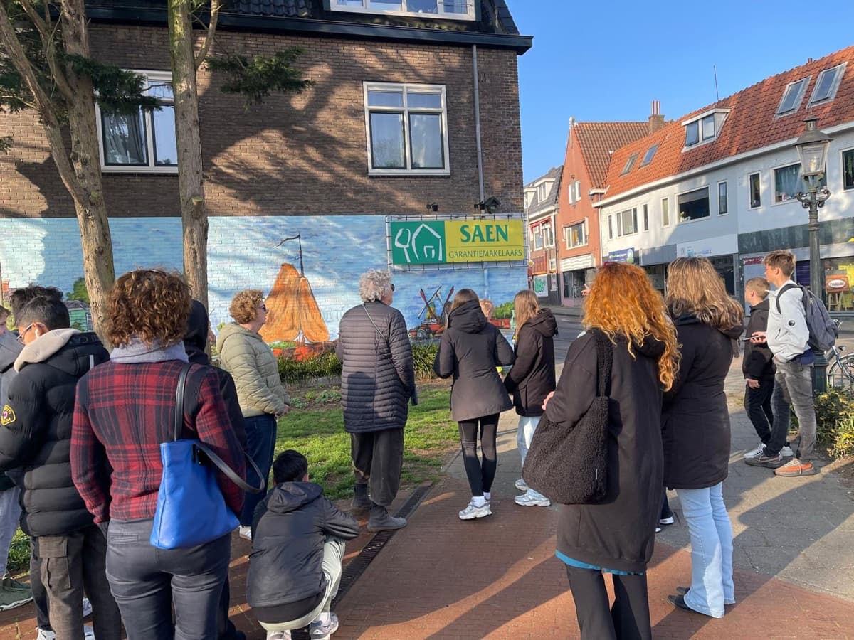 Een klas bekijkt met een gids een muurschildering met een Zaans tafereel